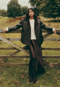 Pruun topeltkarvaline mantel lamba karvaga kraega, valge tekstureeritud kampsun ja põlvpikk tume pitsdetailidega seelik, poseerimas puidust aia ääres.