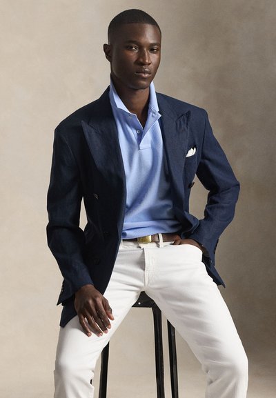 Άνδρας καθισμένος σε σκαμπό, φορώντας μπλε σκούρο σακάκι, ανοιχτό μπλε πόλο πουκάμισο, λευκό παντελόνι και καφέ ζώνη, πάνω σε ουδέτερο φόντο.