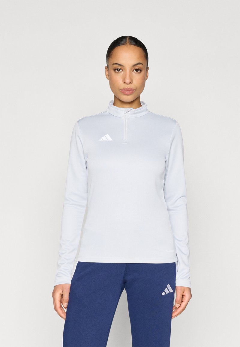 Γυναίκα που φοράει ανοιχτό μπλε μακρυμάνικη μπλούζα με φερμουάρ και σκούρο μπλε παντελόνι, και τα δύο με λευκά λογότυπα Adidas, στέκεται μπροστά σε απλό φόντο.