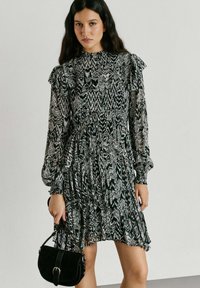 Ženska z dolgimi temnimi lasmi v črno-beli vzorčasti obleki z dolgimi rokavi, ki drži majhno črno torbico ob enobarvni steni.