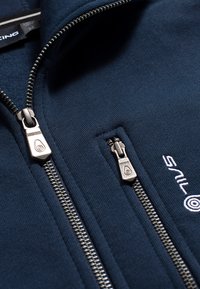 Chaqueta de cremallera azul marino hecha de un tejido suave, con dos cremalleras plateadas, logotipo bordado y detalles texturizados en el cuello.