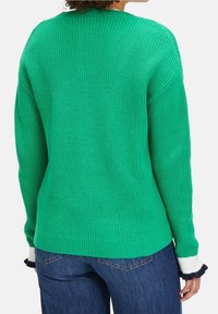 Žena oblečená v jasně zeleném žebrovaném svetru s bílými a černými volánkovými manžetami, v kombinaci s modrými džíny, zobrazená zezadu.