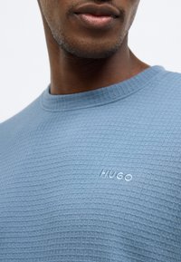 Texturált kék pulóver kerek nyakkivágással és hímzett "HUGO" logóval a bal mellrészen. Puha anyag hálómintával.