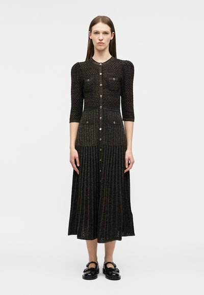 Γυναίκα που στέκεται, φοράει μαύρο υφασμάτινο φόρεμα μεσαίου μήκους με φουσκωτά μανίκια, κουμπιά μπροστά, τέσσερις τσέπες και μαύρα παπούτσια Mary Jane με αγκράφες.