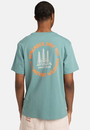 Camiseta de algodón turquesa de manga corta, presenta un gráfico de un árbol blanco y un texto en naranja que dice: "Recuerda, Solo Tú Puedes Prevenir Incendios Forestales."