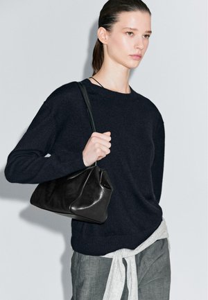Borsa in pelle nera con un design strutturato, tenuta da una persona che indossa un maglione scuro. Pantaloni grigi con una leggera cintura in vita.