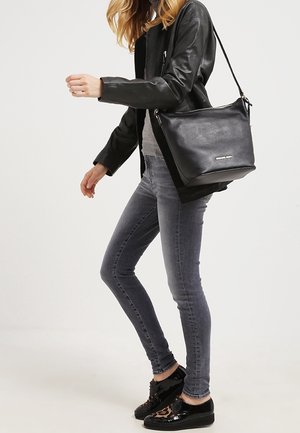 Vrouw draag een zwarte leren jas, grijze strakke jeans, glanzende zwarte sneakers en draagt een zwarte leren crossbody tas over haar schouder.