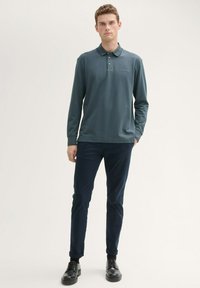 Granatowa koszula polo z długim rękawem w ciemnym odcieniu turkusu, z dwoma guzikami i haftowanym logo, zestawiona z dopasowanymi granatowymi spodniami i czarnymi butami.