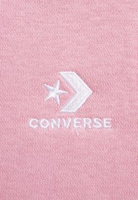 Tela rosa claro con el logotipo "Converse" bordado en blanco, que presenta elementos de diseño como estrellas y chevrons. Textura suave y patrón de punto uniforme.