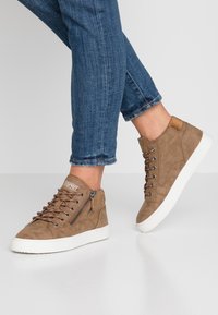 Baskets montantes en daim marron avec semelles en caoutchouc blanc, lacets marron, fermeture éclair latérale et un logo près de la languette.