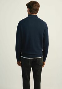 Navy blue sweter z dzianiny z wysokim kołnierzem, ściągaczami na mankietach i u dołu, noszony na białej koszuli, w zestawie z czarnymi dżinsami. Gładka tekstura.