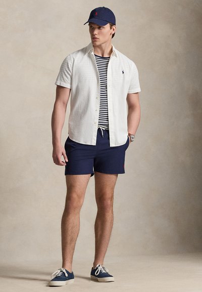 Άνδρας με μπλε σκούφο, λευκό πουκάμισο με κοντά μανίκια και κουμπιά πάνω από ριγέ μπλουζάκι, μπλε σορτς και μπλε αθλητικά παπούτσια, στέκεται με το ένα χέρι στην τσέπη.