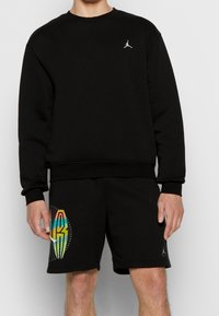 Schwarzes Sweatshirt mit Rundhalsausschnitt und gerippten Bündchen, mit einem kleinen weißen Logo. Kombiniert mit schwarzen Shorts, die ein farbenfrohes Grafikdesign zeigen.