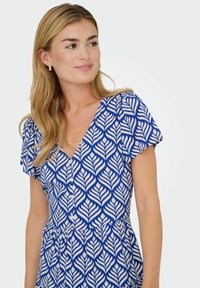 Femme aux longs cheveux blonds portant des boucles d'oreilles en or et une robe à boutons bleu et blanche à motifs, souriant légèrement et regardant vers la droite.
