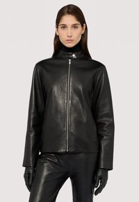 Schwarze Lederjacke mit hohem Kragen, Reißverschluss vorne und langen Ärmeln. Glatte Textur und minimales Zubehör, kombiniert mit schwarzen Lederhandschuhen.