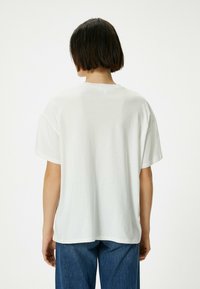 Hvid bomulds T-shirt med en afslappet pasform, korte ærmer og rund halsudskæring, vist fra ryggen mod en ensfarvet baggrund.