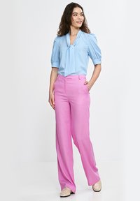 Kobieta stoi z ręką w kieszeni, ubrana w jasnoniebieską bluzkę z kokardą oraz różowe, szerokie spodnie z wysokim stanem i beżowe półbuty.