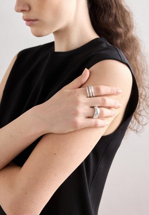 Anelli d'argento indossati sulle dita. La canotta nera senza maniche del modello presenta un collo rotondo. Il tono della pelle è chiaro con lentiggini.