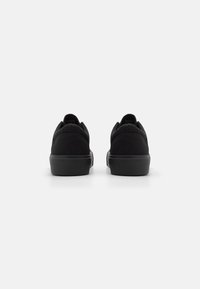 Black canvas sneakers viewed from the back, featuring a textured rubber sole and a low-profile design with padded collar accents.