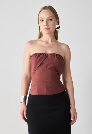 Top fără bretele din material roșu închis, cu detalii plisate și design unghiular, asortat cu o fustă neagră. Brățară din argint purtată pe încheietura mâinii.