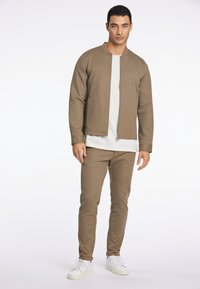 Giacca bomber a maglia marrone sopra una t-shirt bianca, abbinata a pantaloni marroni coordinati e sneakers bianche. Texture liscia, design minimalista.