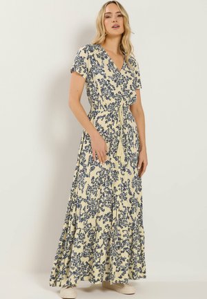 Vrouw draagt een crèmekleurige maxi-jurk met korte mouwen, marineblauwe bloemenprint en een taillekoord, staand tegen een eenvoudige lichte achtergrond.