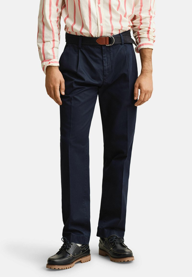 Homme portant un pantalon plissé bleu marine avec ceinture noire, une chemise rayée rose et blanche, et des chaussures en cuir noires à lacets avec semelles marron.