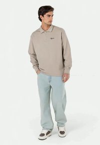 Beige long-sleeve polo shirt with a zip collar and embroidered logo, paired with light blue loose-fit jeans and white sneakers with brown accents.