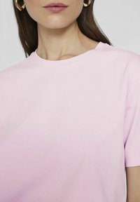 En närbild av en kvinnas halslinje och axlar som bär en enkel rosa t-shirt, med stora guldörhängen.