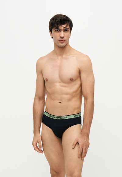 Jeune homme aux cheveux courts et foncés posant torse nu, portant un slip noir Emporio Armani avec une ceinture verte sur un fond clair uni.