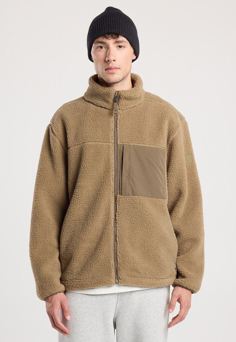Bruine fleece jack met een hoge kraag en volledige rits; heeft een borstzak in een donkerdere tint en geribbelde mouwen. Draagt met een zwarte beanie.