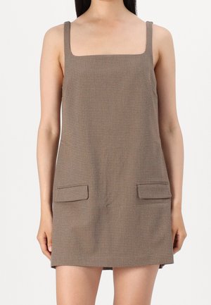 Mujer con un vestido mini marrón sin mangas de cuadros, con escote cuadrado y dos bolsillos con solapa en la parte delantera.