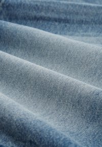 Lähellä näkyvää vaaleansinistä farkkukangasta, jossa on viistoon kudottu rakenne ja pehmeät taitteet.