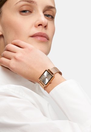 Roze-gouden vierkante horloge met een mesh-band, zilveren wijzerplaat en minimalistisch ontwerp. Draagt om een pols, wat het stijlvolle profiel benadrukt.