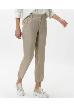Vrouw met beige taps toelopende broek en witte sneakers, staand met één been iets naar voren op een witte achtergrond.