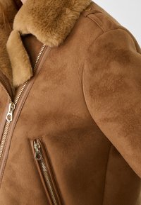 Giacca in camoscio marrone con colletto in pelliccia sintetica morbida, tasche con zip e dettagli in oro, che mettono in risalto la sua morbidezza e il design caldo.