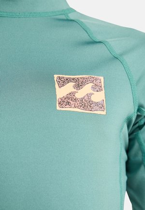 Nahaufnahme von türkisfarbenem Stoff mit einem kleinen Aufnäher, der auf der Brust ein stilisiertes Wellenmuster in Pink und Schwarz zeigt.