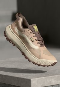 Beige atletik sko med en strukturert såle, lagdelt overdesig og brune detaljer. Funksjoner inkluderer en polstret krage og logo detaljer.