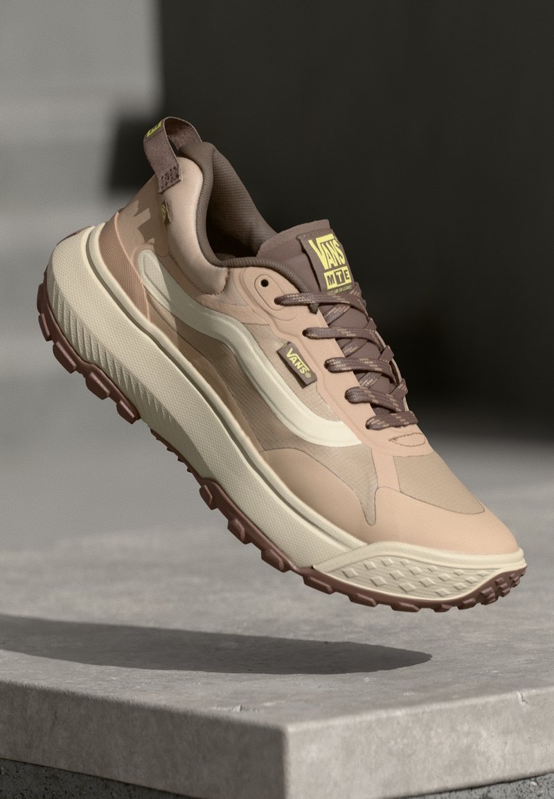 Beige atletik sko med en strukturert såle, lagdelt overdesig og brune detaljer. Funksjoner inkluderer en polstret krage og logo detaljer.