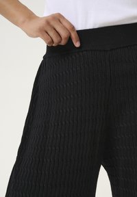 Pantalon noir côtelé en tissu texturé, doté d'une large ceinture élastique, tenus à la taille, avec des lignes verticales détaillées.