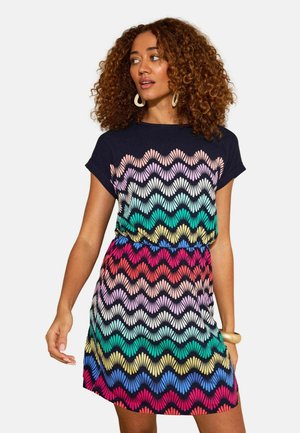 Göndör hajú nő, akinek rövid ujjú, többszínű, hullámos mintás ruhája és nagy karika fülbevalói vannak, oldalra néz.