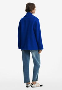 Manteau en laine bleu vif, texturé, avec un col en V, une coupe ample et des épaules tombantes, associé à un jean léger bleu clair et des chaussures blanches.