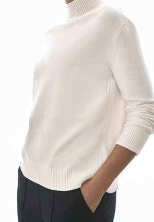 Osoba ubrana w biały prążkowany golf i czarne spodnie, z jedną ręką w kieszeni, na białym tle.