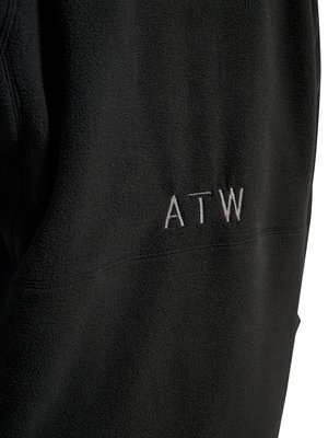 Sort fleece-stof med hvide broderede store bogstaver "ATW" på bagsiden, der viser tekstureret materiale og syningsdetaljer.