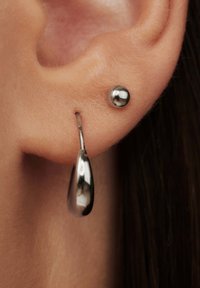 Silberne Ohrringe mit einer Tropfenform und einer kleinen Kugel, mit glatter Oberfläche. Hängend an einem dünnen Ring, der im Kontrast zum Hautton steht.