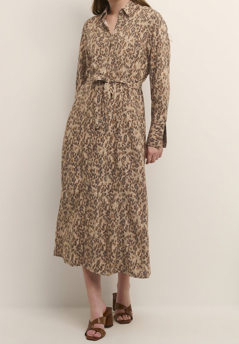 Robe à manches longues et à boutons dans un imprimé léopard beige et marron. Présente une taille ceinturée et une coupe mi-longue. Texture de tissu lisse.