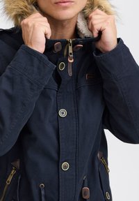 Mørkeblå jakke med faux fur-foret hætte, guld lynlås, brune læderdetaljer, trykknap- og lynlås-lukning samt sidelommer. Struktureret stof.