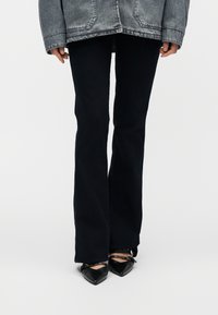 Personne portant un pantalon noir évasé, des chaussures noires à bout pointu avec des lanières, et une veste en denim grise, se tenant devant un fond uni.