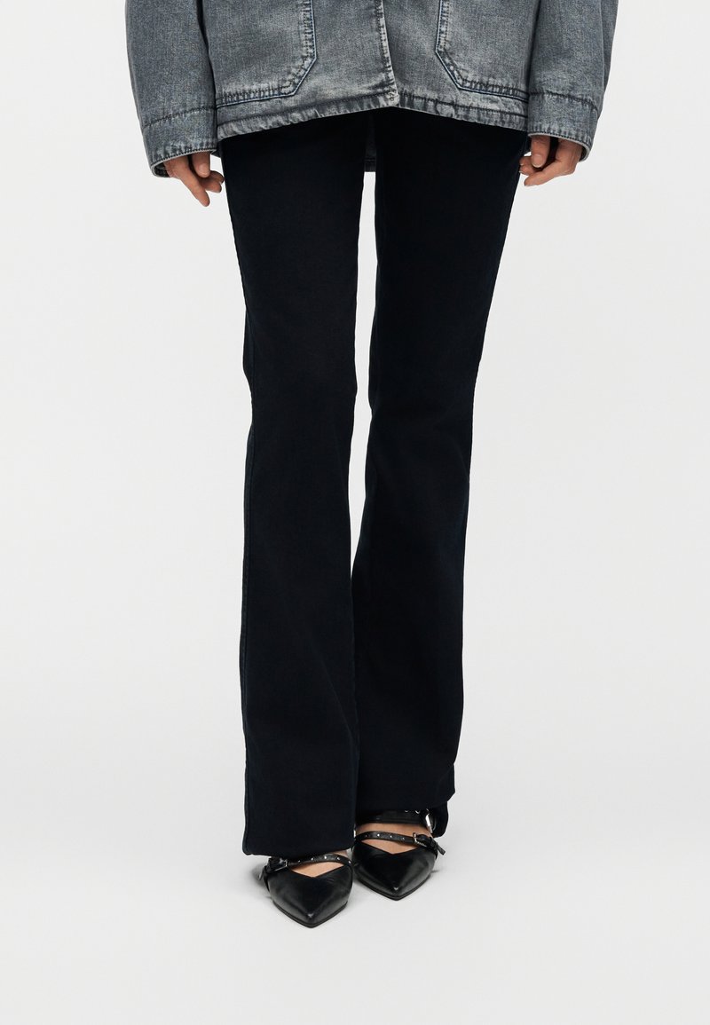 Personne portant un pantalon noir évasé, des chaussures noires à bout pointu avec des lanières, et une veste en denim grise, se tenant devant un fond uni.