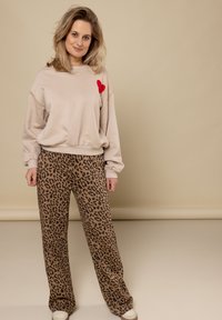 Beige sweatshirt met rode hartdetail, gecombineerd met brede bruine broek met luipaardprint. Model staat zelfverzekerd tegen een neutrale achtergrond.